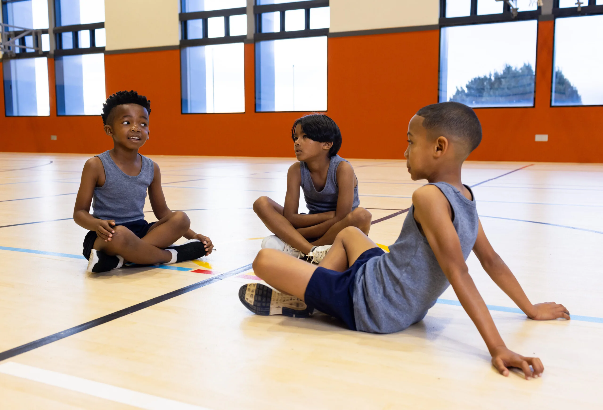 dans la salle de sport de l ecole trois garcons multiraciaux assis sur le sol en uniforme de sport se parlent scaled